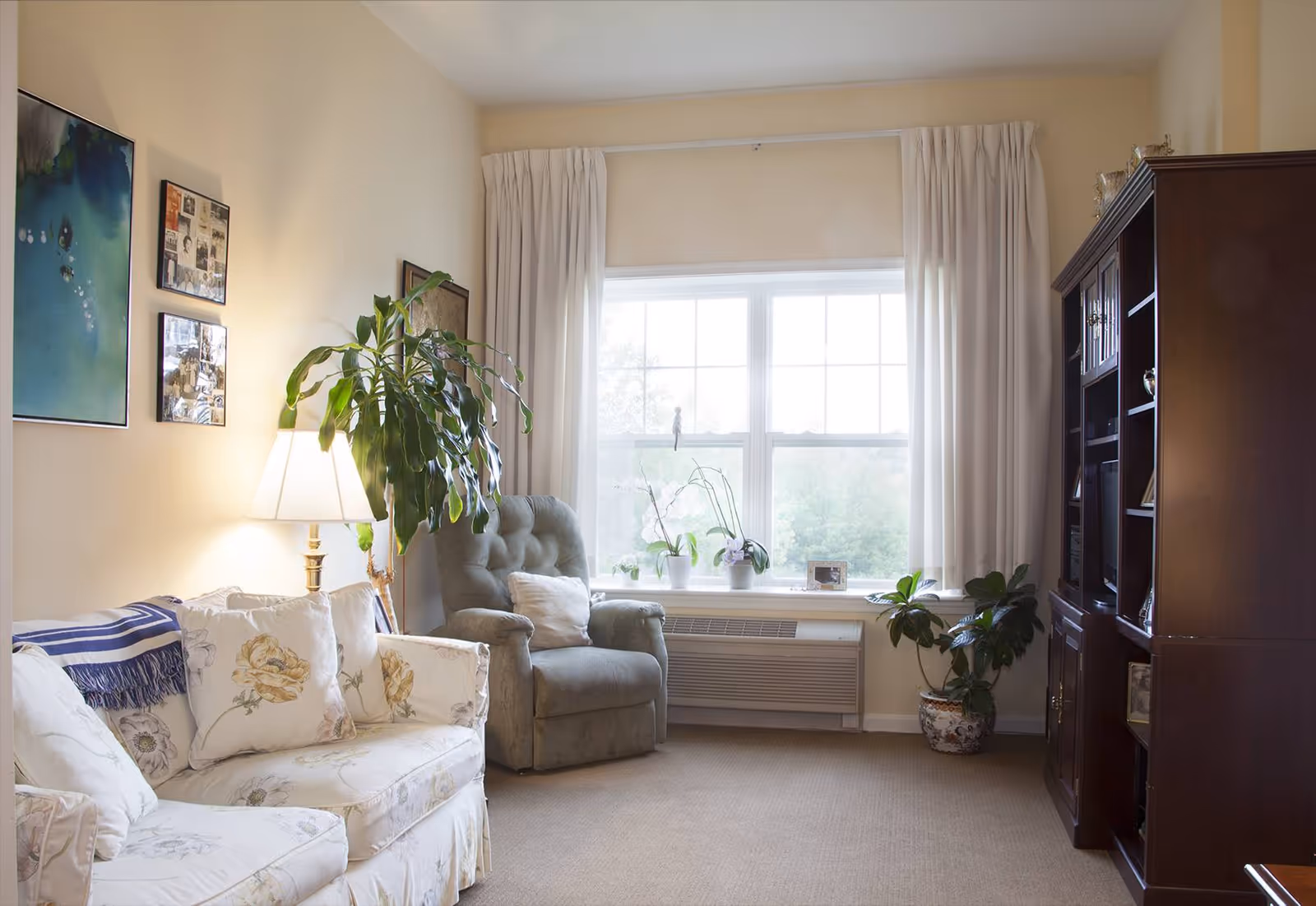A cozy living room with a floral-patterned sofa, a gray armchair, a floor lamp, several potted plants, and a large wooden bookshelf. The room has a large window with white curtains letting in natural light.