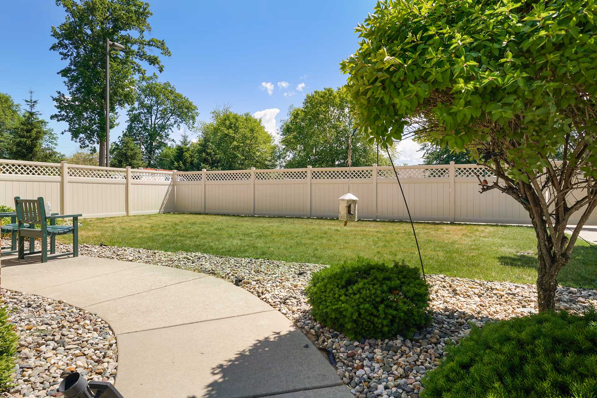 A fenced outdoor garden area with a concrete pathway, green grass, small bushes, a tree, and a green bench under a clear blue sky.