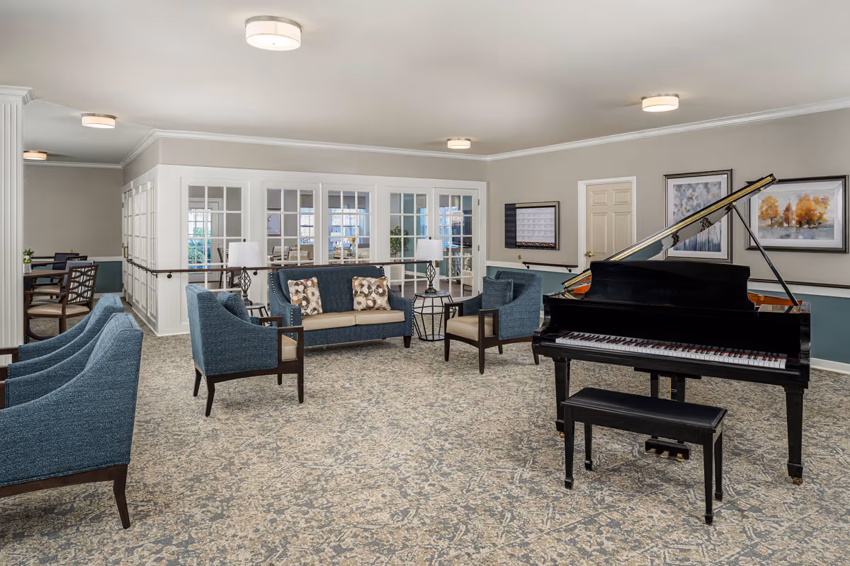 A spacious and well-lit common area in a senior living facility featuring a black grand piano with a matching bench, several blue upholstered armchairs, a blue sofa with patterned cushions, two side tables with lamps, and framed artwork on the walls. The room has a patterned carpet and large windows with white frames in the background.