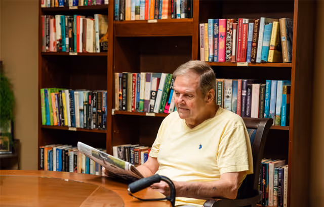 An elderly man wearing a yellow shirt is sitting at a wooden table in a library or reading room, reading a newspaper. Behind him are bookshelves filled with various books. A black cane rests on the table in front of him.