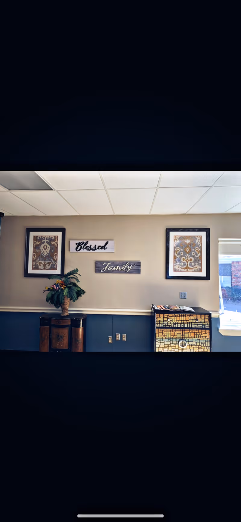 Interior wall of a room with two framed decorative artworks, two wooden signs reading 'Blessed' and 'Family', a small table with a potted plant, and a mosaic-style cabinet near a window showing a partial outdoor view.
