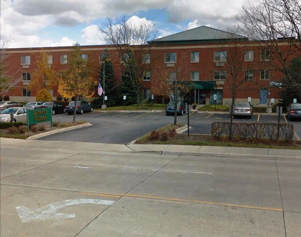 Exterior front view of Liberty Village Senior Apartments, a three-story brick building with several windows. There is a parking lot with cars in front, some trees with autumn foliage, and an American flag near the entrance. The sky is partly cloudy.