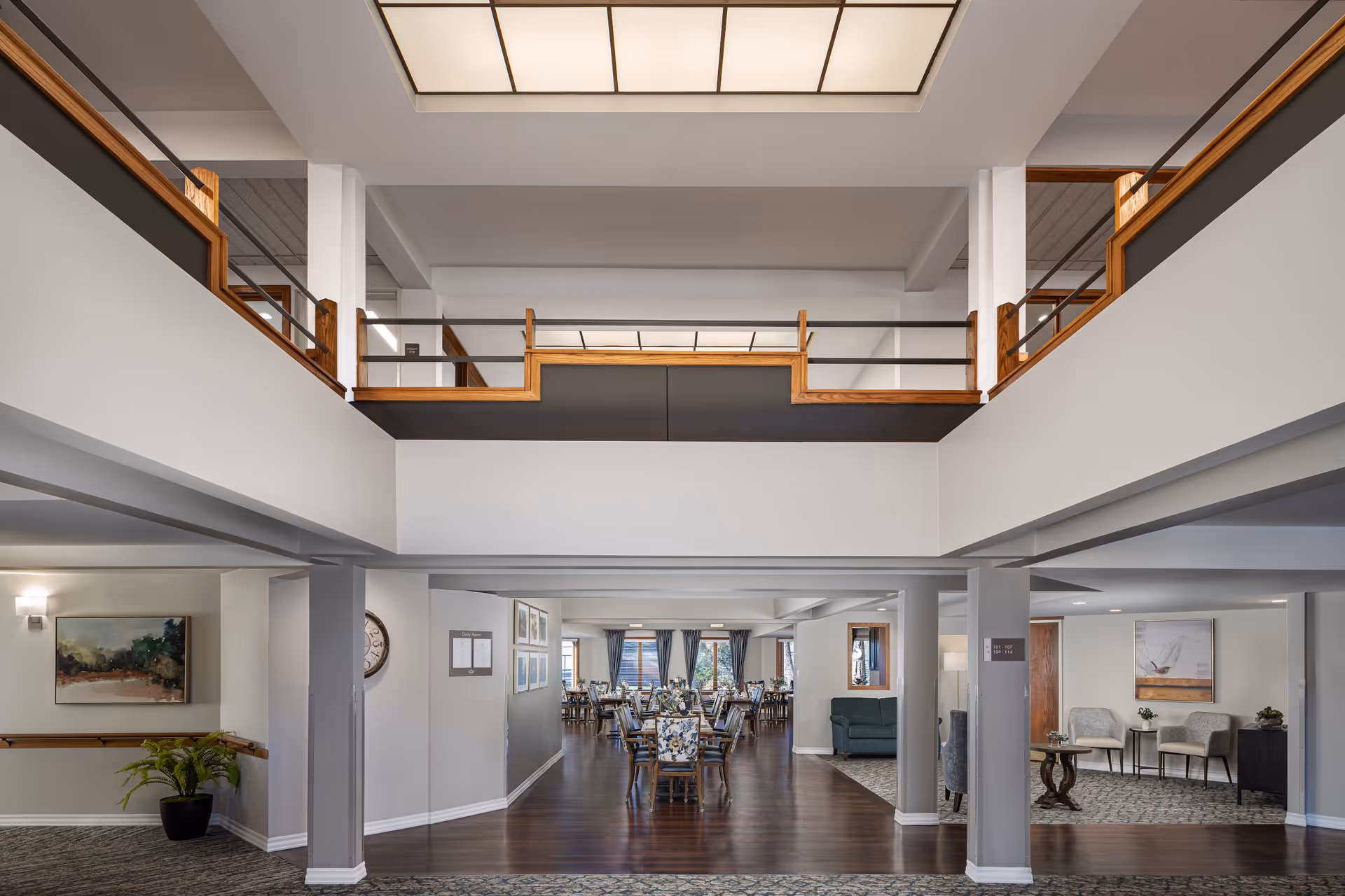 Two-story interior common area of a senior living community with a central dining room, seating areas, and an upper mezzanine balcony.
