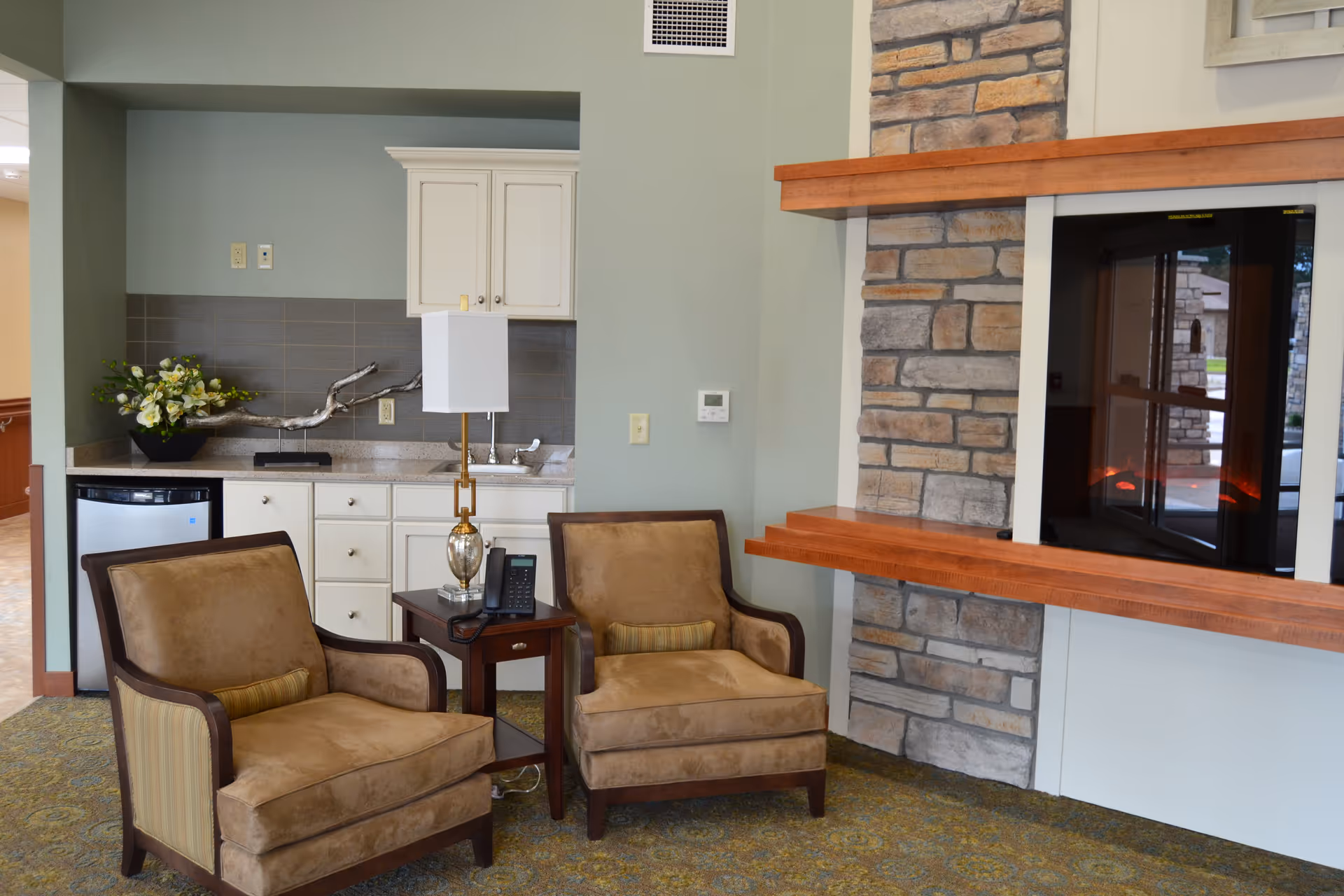 A cozy interior seating area with two cushioned armchairs and a small wooden side table with a lamp and a phone. Behind the chairs is a kitchenette with white cabinets, a small refrigerator, a sink, and a decorative flower arrangement. To the right is a stone fireplace with a wooden mantel.