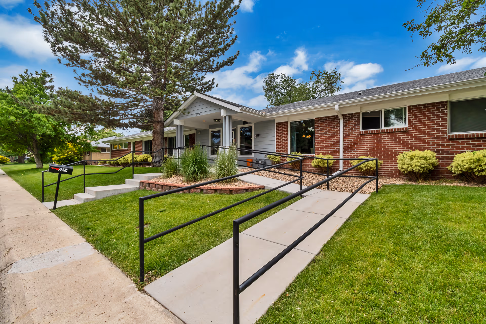 Front exterior of a single-story brick senior living building with an accessible ramp, steps, and landscaped lawn.