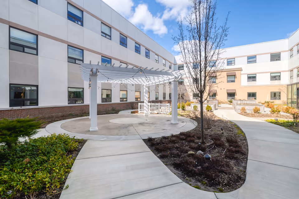 Outdoor courtyard area of a senior living facility with a white pergola structure, paved walkways, landscaped garden beds, and a three-story building surrounding the courtyard under a partly cloudy blue sky.