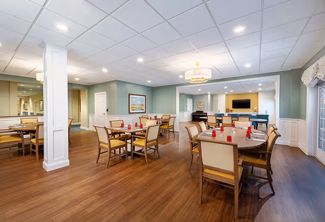 A spacious senior living dining room with several wooden tables and chairs arranged neatly. Each table has red napkin holders and place settings. The room features wood flooring, light green walls with white wainscoting, a ceiling with recessed lighting, and a chandelier. There is a large mirror on one wall and a flat-screen TV mounted on a yellow accent wall in the background.