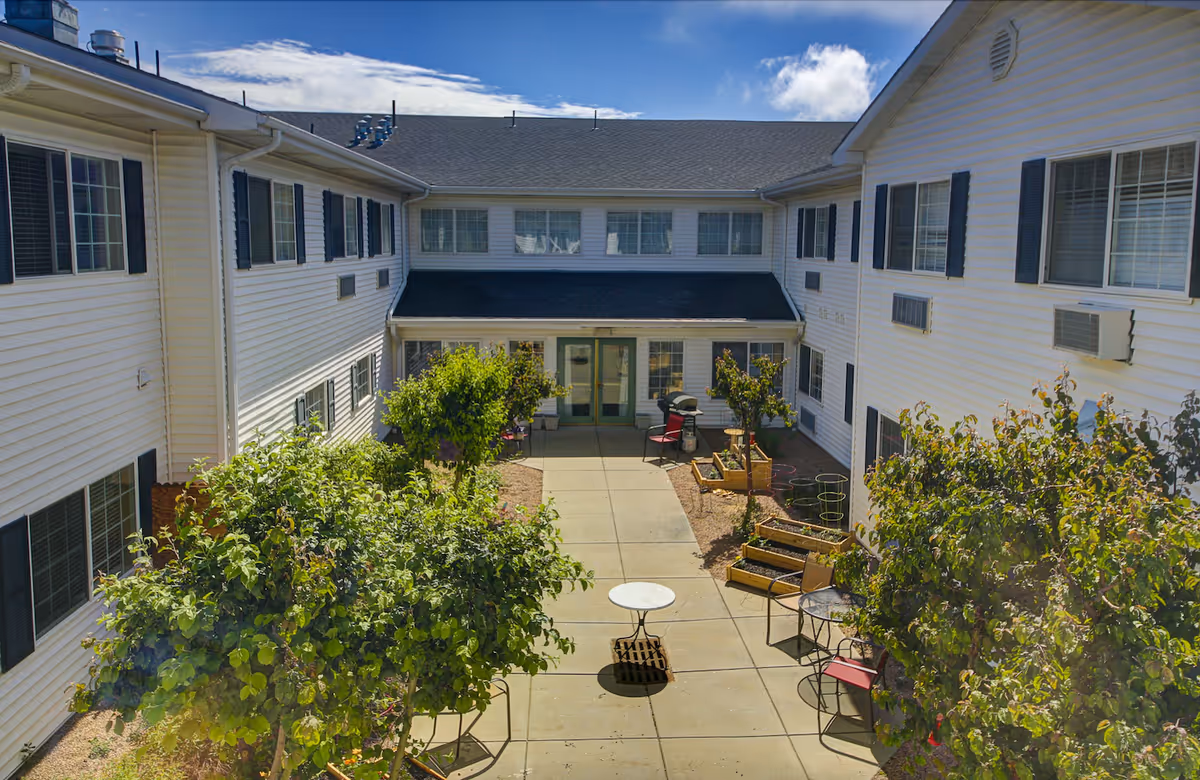 Sunlit central courtyard of a two-story assisted living building with a paved walkway, patio tables, chairs and raised planters surrounded by white siding.