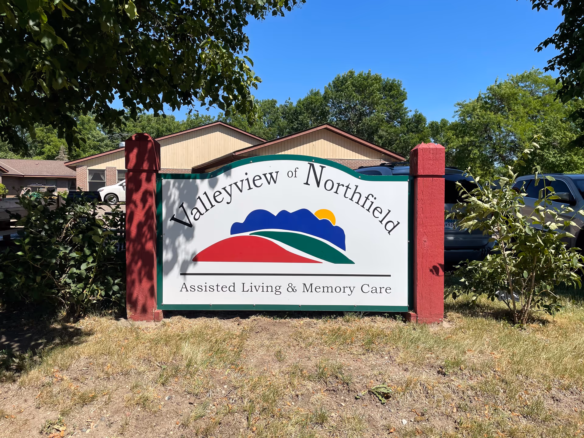 Outdoor sign for Valleyview of Northfield Assisted Living & Memory Care with a colorful logo of hills and a sun, set in front of a building and surrounded by trees and parked cars under a clear blue sky.