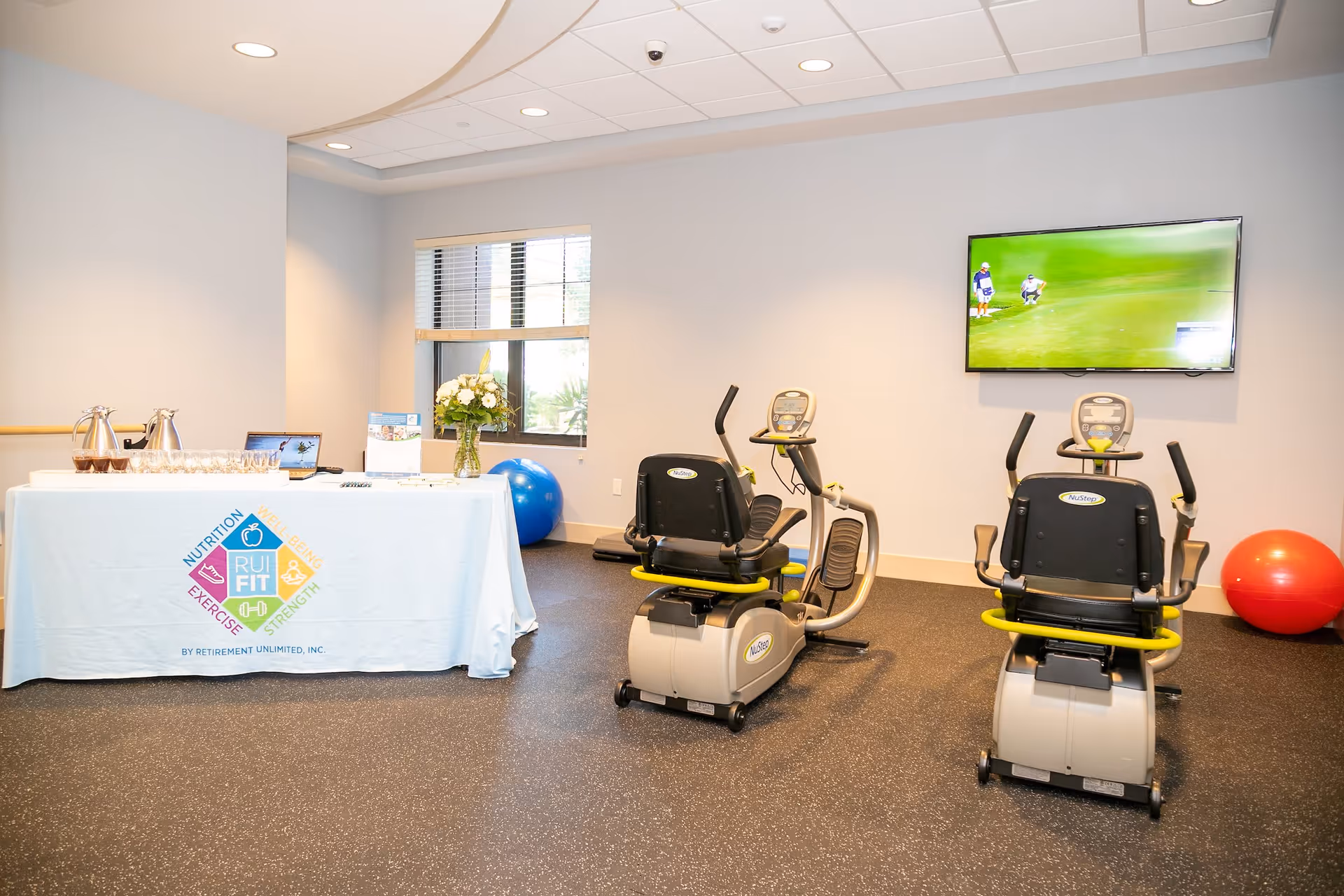 A fitness room with two stationary exercise bikes facing a wall-mounted TV showing a golf game. To the left, there is a table covered with a white cloth displaying the logo and text 'RUI FIT' along with words Nutrition, Wellness, Exercise, and Strength. The table holds a laptop, a vase with flowers, and some glassware. There is a blue exercise ball near the window and a red exercise ball in the corner.