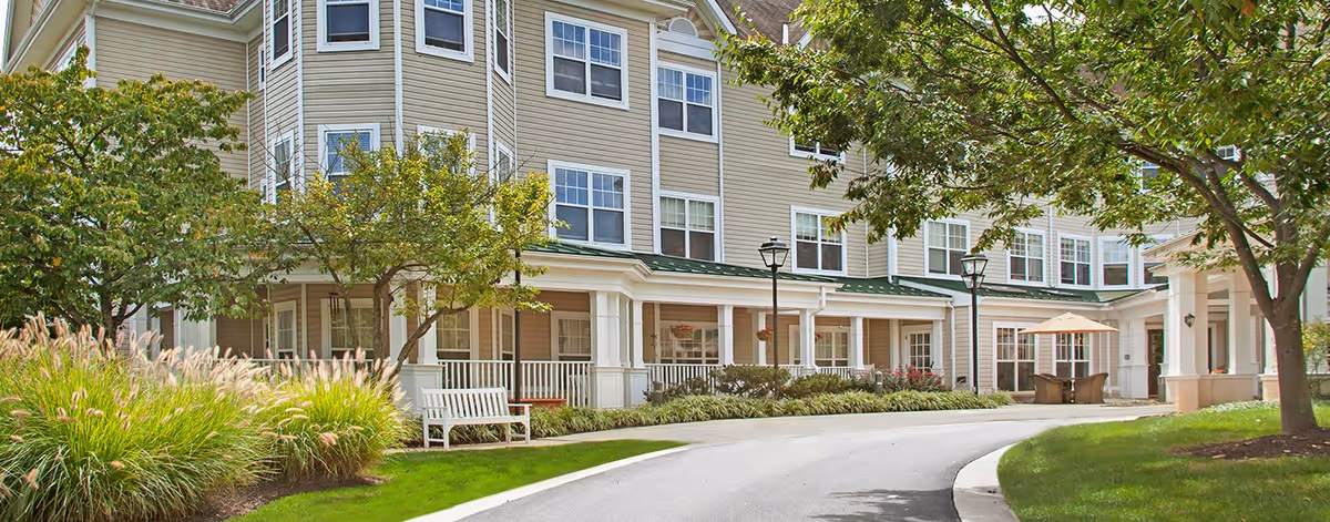 Exterior front of a multi-story senior living building with a curved driveway, benches, trees, and landscaped grounds.