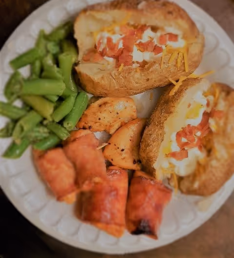 A plate with two loaded baked potatoes topped with sour cream, cheddar and bacon, served with green beans and bacon-wrapped items.