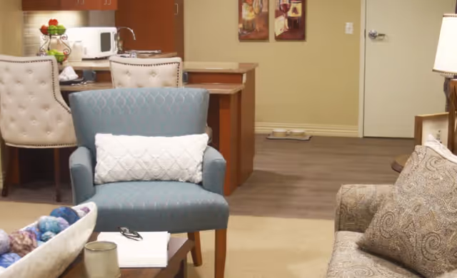 A cozy living area with a blue armchair featuring a white textured pillow, a patterned sofa with matching pillows, and a wooden coffee table holding a candle, a pair of glasses, and a book. In the background, there is a kitchen area with a microwave, a sink, and two beige upholstered chairs at a counter. The floor transitions from carpet to wood, and there is a door and two framed pictures on the wall.