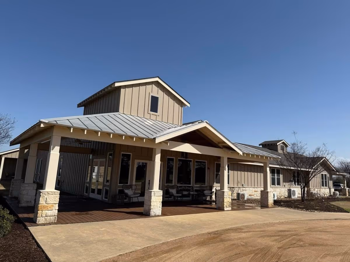 Beige single-story building with a covered porch entrance supported by stone pillars and a metal roof under a clear blue sky.