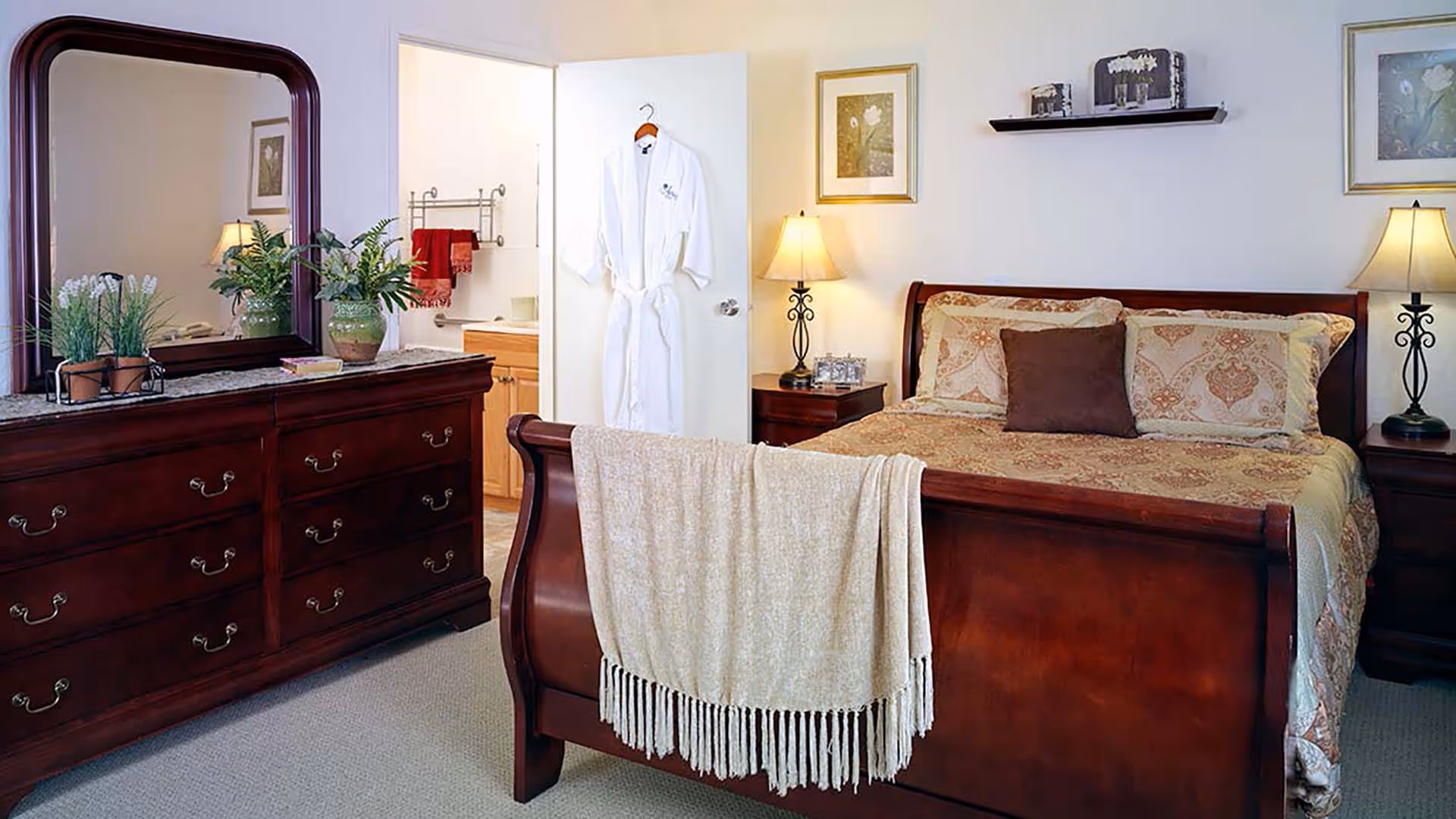 A cozy bedroom with a wooden sleigh bed covered in patterned bedding and a brown pillow. A beige throw blanket is draped over the footboard. On either side of the bed are matching wooden nightstands with lamps. A wooden dresser with a large mirror and potted plants is on the left. The door to an adjoining bathroom is open, showing a white robe hanging on the door and red towels inside the bathroom.