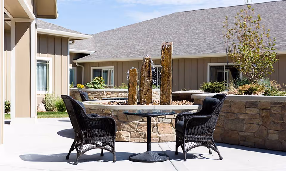 Outdoor courtyard with wicker chairs and a table around a stone fountain in front of a single-story building.