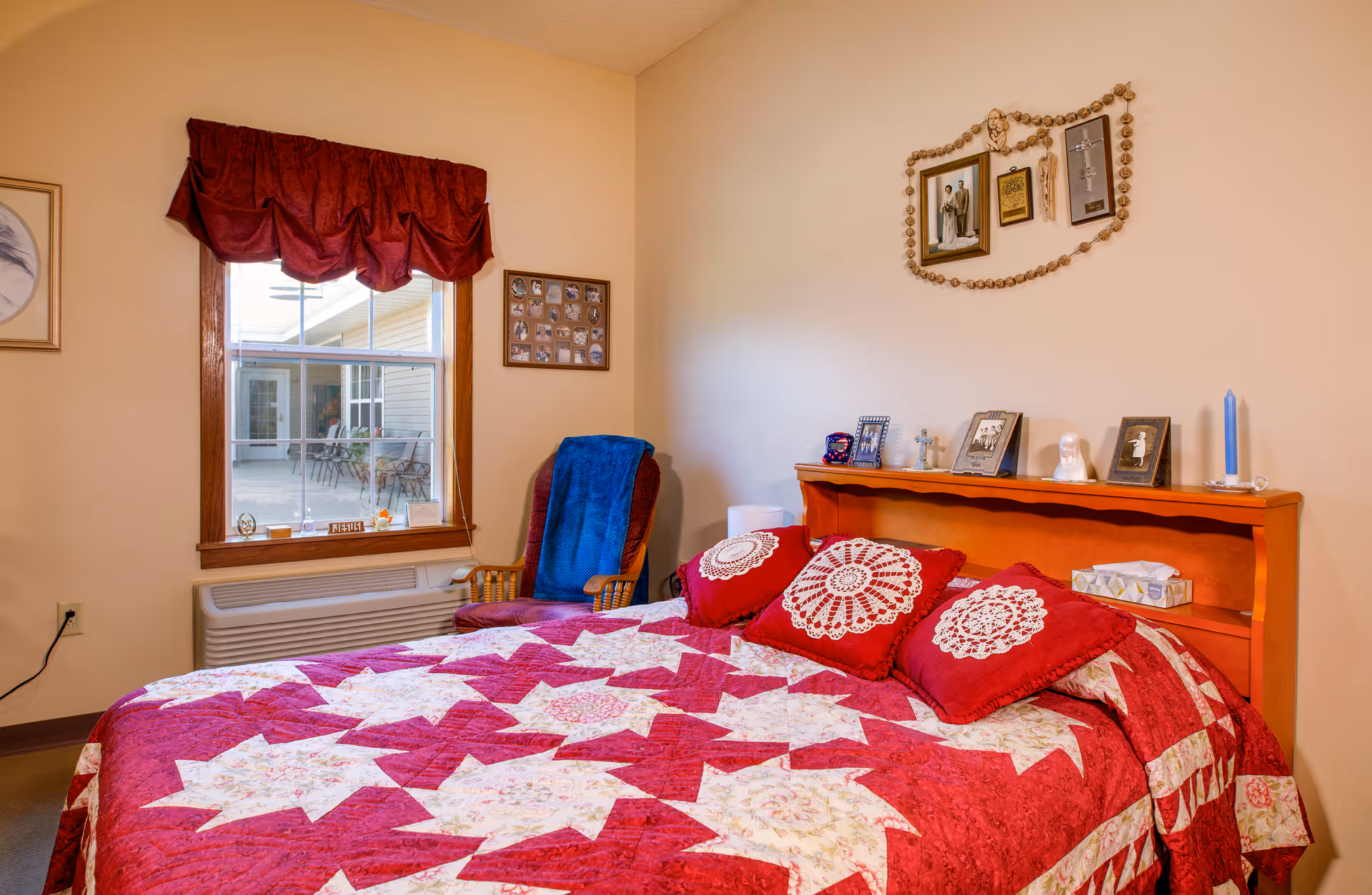 Well-lit bedroom with a red patterned quilted bed, wooden headboard topped with framed photos and decor, a window with a valance and a chair beside it.