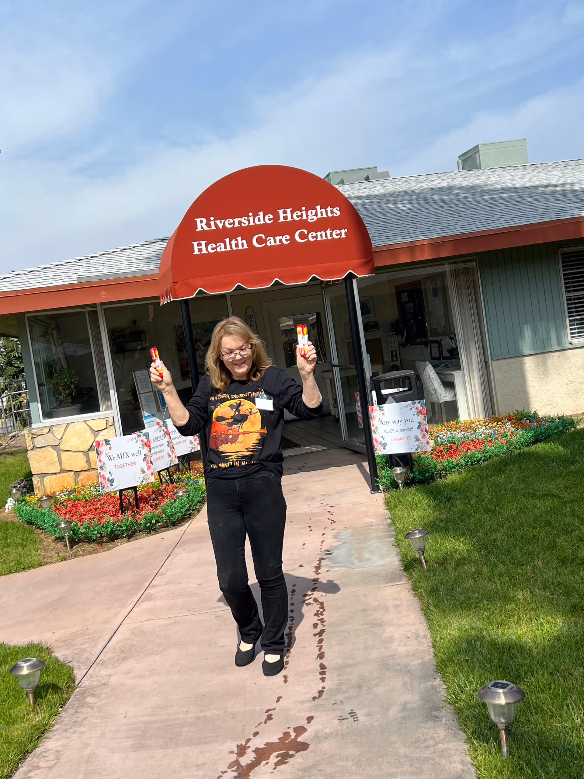 A woman standing on a sidewalk in front of the entrance to Riverside Heights Health Care Center, holding two popsicles and smiling. The entrance has a red awning with the facility's name, and there are flower beds and signs on either side of the walkway.