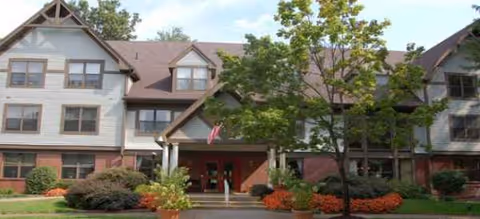 Exterior view of Brookdale Gables Farmington entrance with trees, shrubs, and a red doorway.