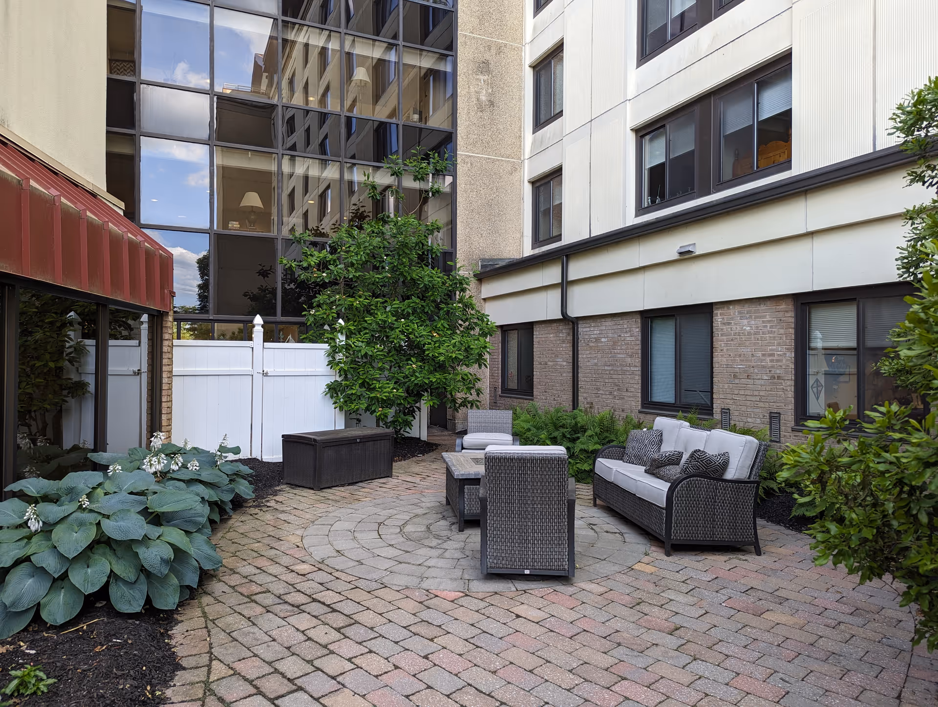 Outdoor patio area with cushioned wicker seating including a sofa, chairs, and a coffee table, surrounded by plants and trees. The patio is paved with bricks and enclosed by the building walls with windows reflecting the sky.