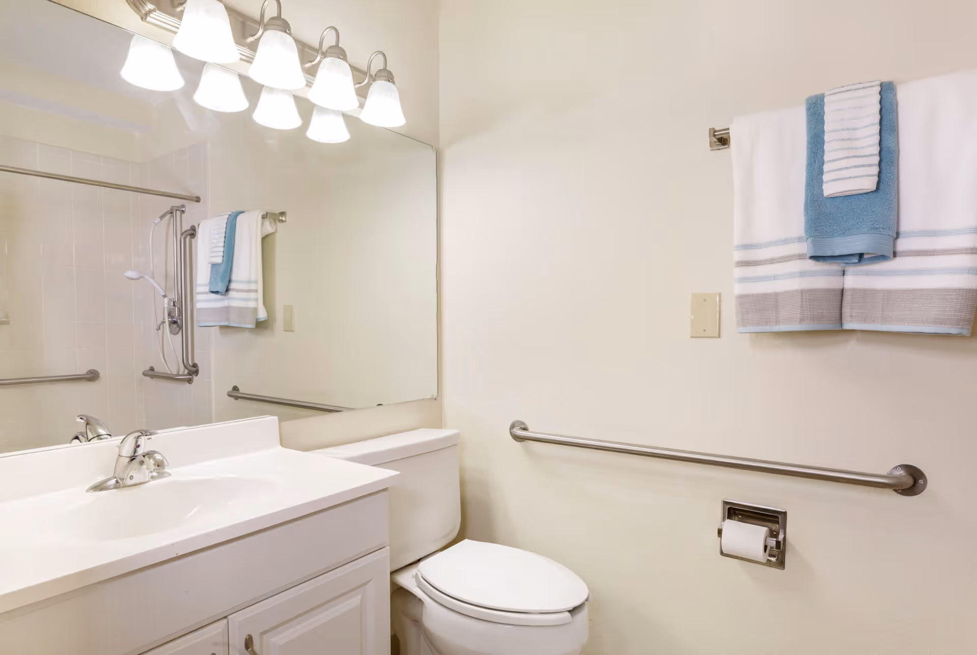 A clean and bright bathroom featuring a white sink with a silver faucet, a large mirror above the sink, a white toilet, and a towel rack with neatly folded white and blue towels. There are safety grab bars installed near the toilet and in the shower area, which is partially visible with a showerhead and curtain rod.