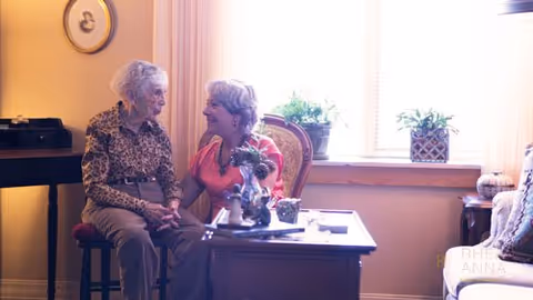 Two elderly women sitting and holding hands in a warmly lit living room with a window, plants on the windowsill, and a table with decorative items in front of them.