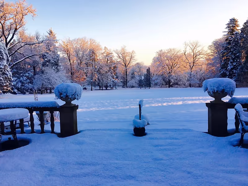 Snow-covered outdoor patio area with tables, chairs, and large planters, overlooking a wide open snow-covered field and trees with snow on their branches during sunrise or sunset.