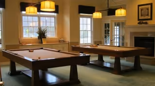 A spacious game room with two pool tables, large windows letting in natural light, hanging pendant lights above the tables, a fireplace on the right wall, and a framed picture above the fireplace.
