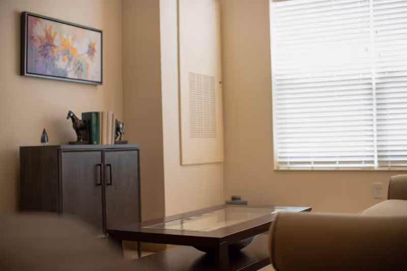 A cozy living room area with a glass-top coffee table, beige armchairs, a dark wooden cabinet with decorative items and books, a colorful abstract painting on the wall, and a window with white blinds letting in natural light.