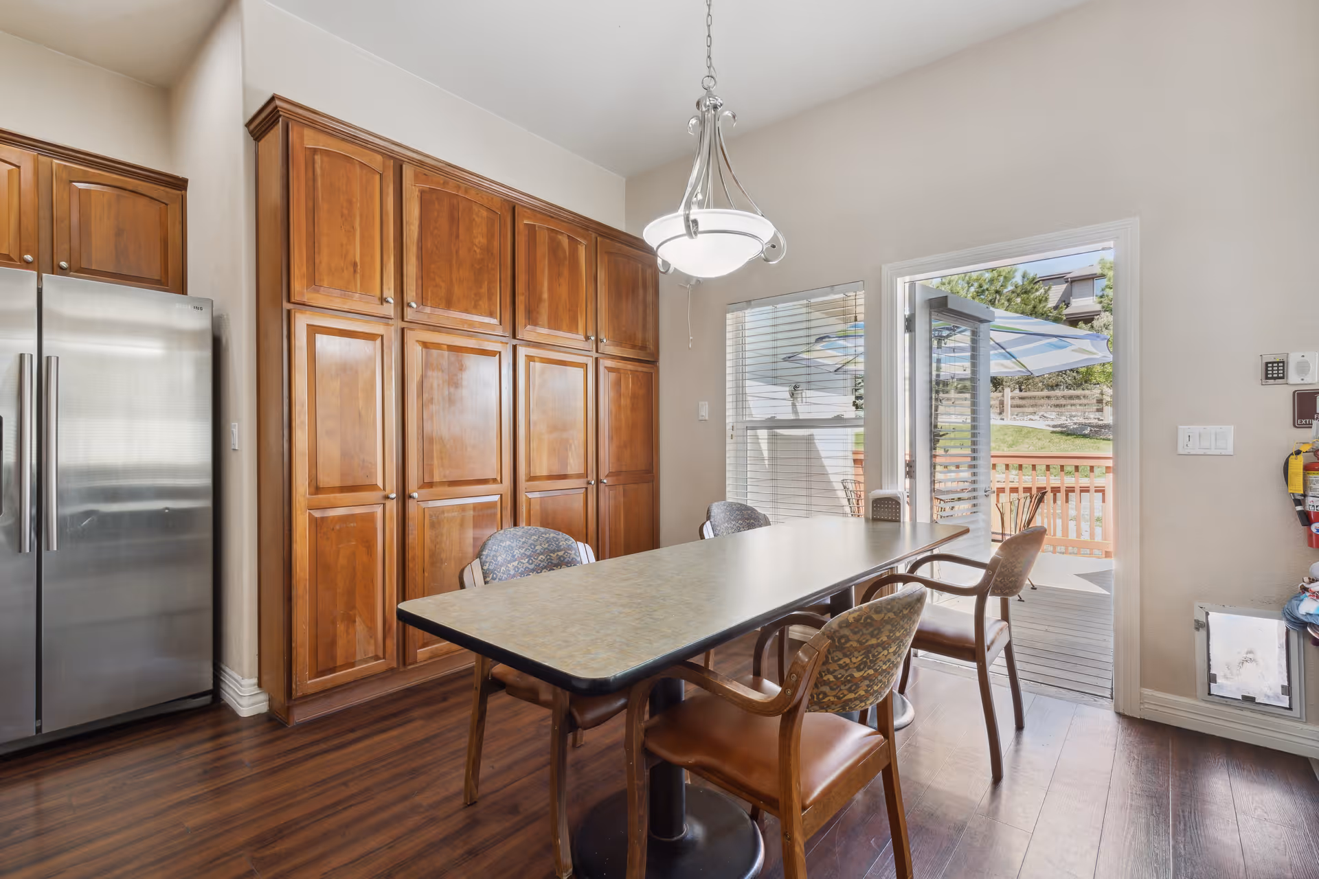 Dining area with a long table and chairs, tall wooden cabinets, a stainless steel refrigerator, and a door opening onto an outdoor deck.