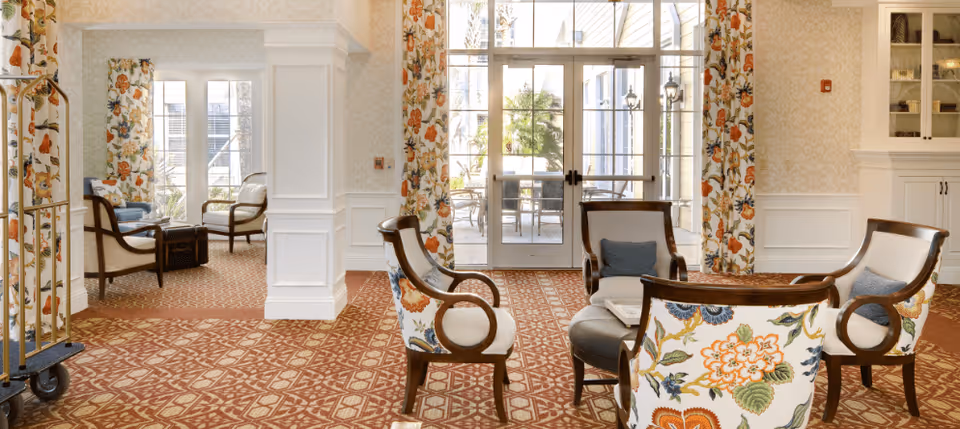 A bright and elegant sitting area in a senior living facility with floral-patterned chairs arranged around a small round table. The room features large windows and glass doors leading to an outdoor patio, floral curtains, and a patterned carpet. There is a luggage cart on the left side and built-in cabinetry on the right.