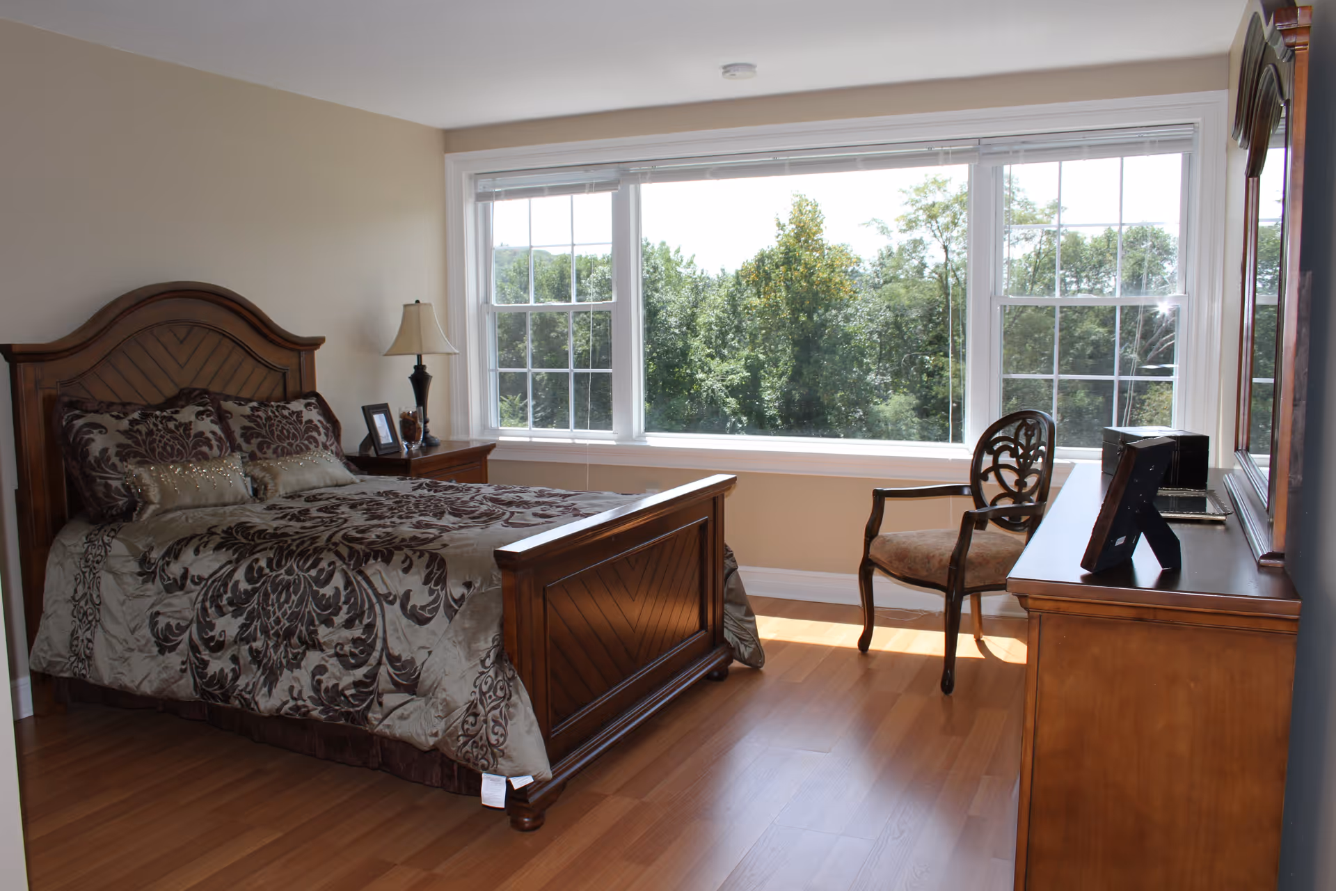 A bedroom with a large wooden bed featuring a patterned bedspread and pillows. Next to the bed is a wooden nightstand with a lamp and picture frames. Across from the bed is a wooden dresser with a mirror and a decorative chair. A large window behind the chair shows a view of green trees outside.