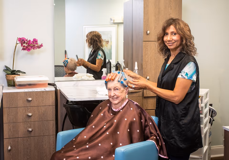 An elderly woman with hair rollers sits in a blue salon chair wearing a brown polka dot cape while a hairstylist stands beside her smiling and adjusting the rollers. The setting is a hair salon area with wooden cabinets, a large mirror, and a potted orchid on the counter.