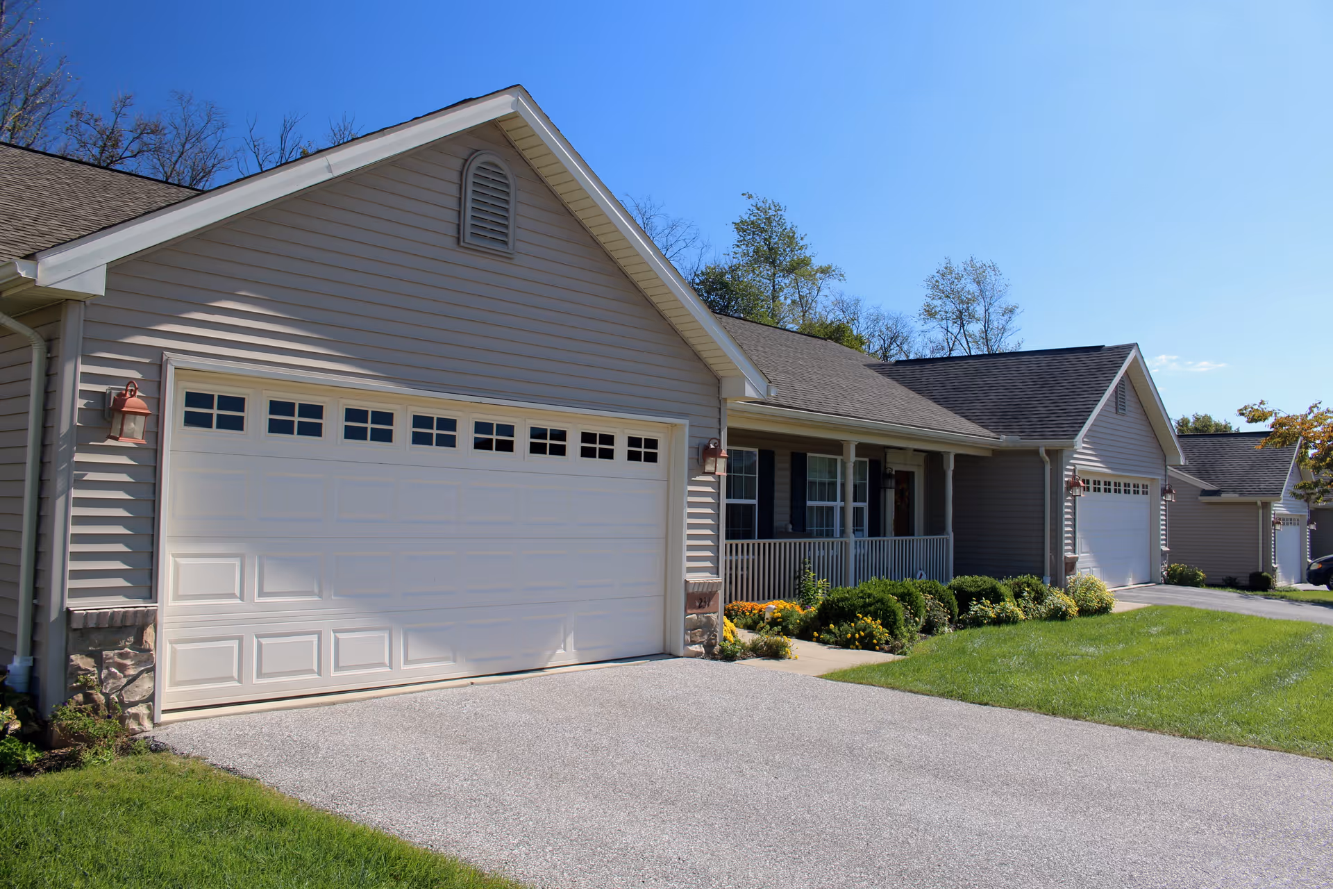 Exterior view of a single-story residential building with beige siding, a white garage door, a small front porch with white railing, and a well-maintained lawn and garden under a clear blue sky.