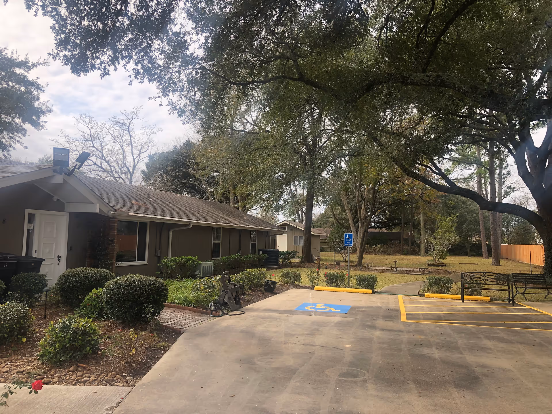 Outdoor view of a single-story assisted living facility building with bushes and trees surrounding it. There is a handicapped parking space in the foreground and a bench near a pathway leading into a grassy area with more trees.