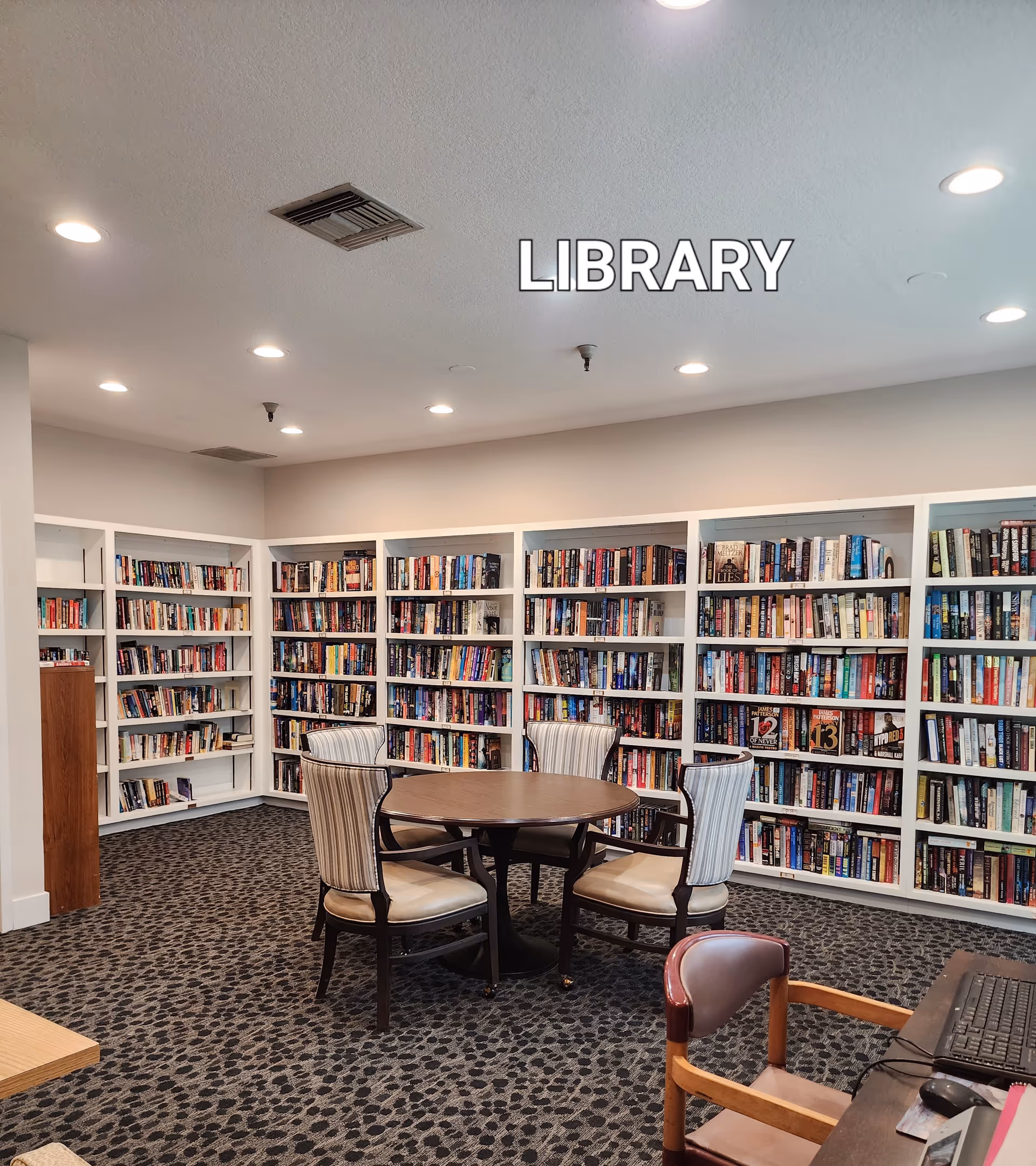 Interior view of a library room with white bookshelves filled with books along the walls. In the center, there is a round wooden table surrounded by four upholstered chairs with striped fabric. The carpet has a patterned design, and there is a desk with a computer and chair in the foreground. The ceiling has recessed lighting and an air vent. The word 'LIBRARY' is displayed prominently on the wall above the bookshelves.