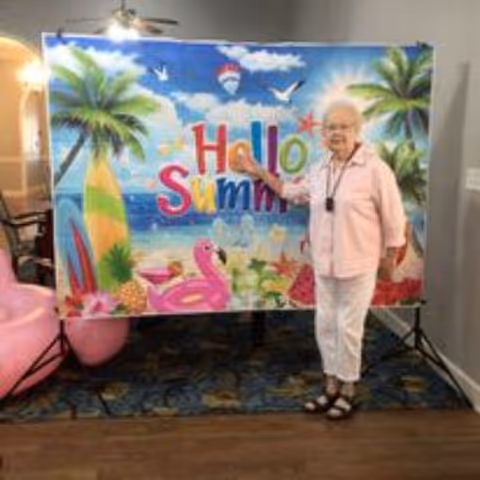 An elderly woman standing indoors next to a colorful summer-themed backdrop that says 'Hello Summer' with images of palm trees, a flamingo, and a beach scene.