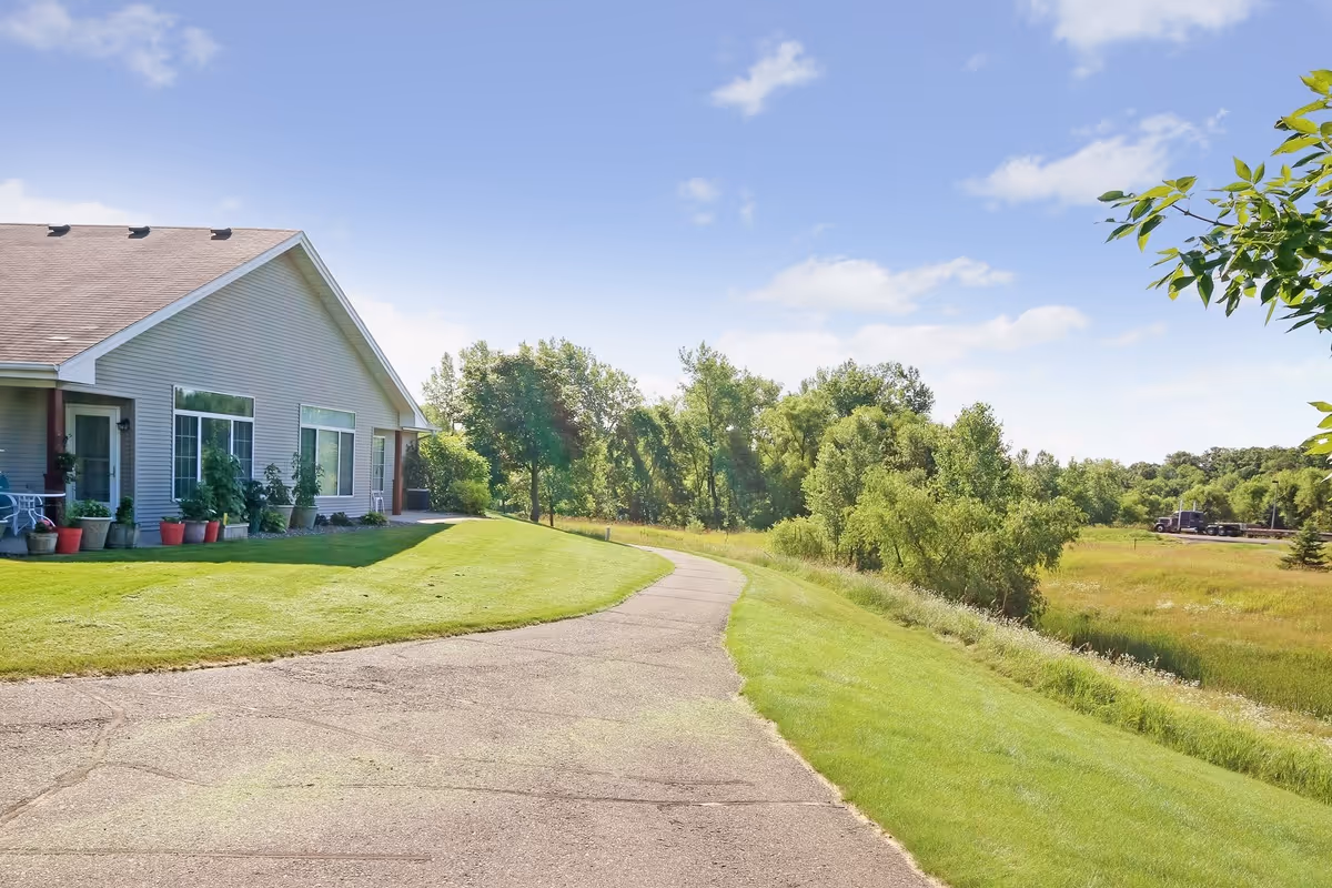 A single-story building with beige siding and a brown roof next to a paved pathway surrounded by green grass and trees under a blue sky with scattered clouds.