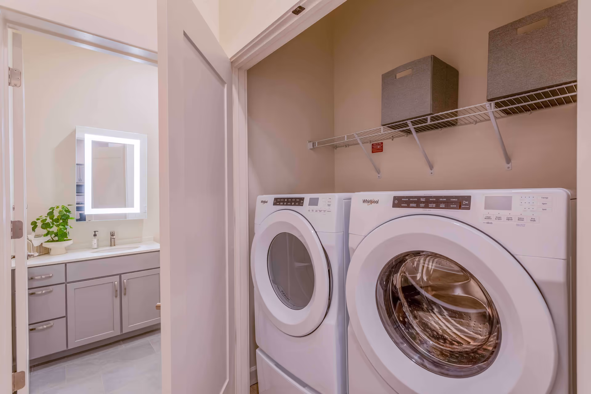 Laundry area with a white Whirlpool front-loading washing machine and dryer side by side. Above them is a white wire shelf holding two gray storage bins. To the left, an open door reveals a bathroom with a lighted mirror, a countertop with a plant, soap dispenser, and tissue box, and gray cabinets below.