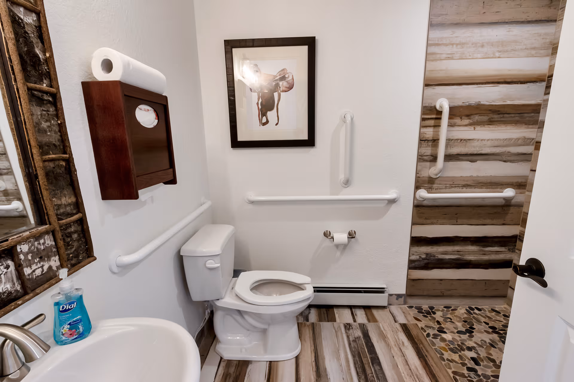Small accessible bathroom with a toilet, sink, multiple grab bars, wood-look flooring and framed artwork.