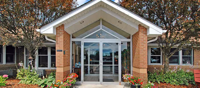 Front entrance of a brick building with a peaked roof over the doorway, glass double doors, and landscaped flower beds with green plants and colorful flowers on either side. Trees with autumn foliage flank the entrance.