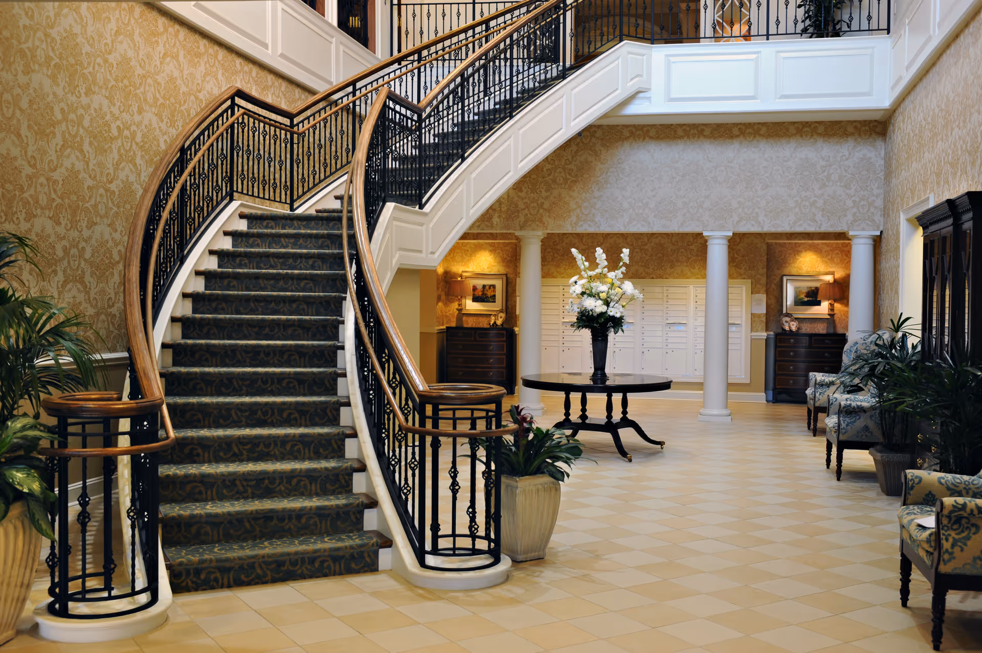 Elegant interior of a senior living facility featuring a grand curved staircase with ornate black railings and wooden handrails. The floor is tiled in a light beige pattern, and the walls are decorated with gold damask wallpaper. In the center, there is a round table with a large floral arrangement. The space includes potted plants, upholstered chairs with floral patterns, white columns, and a set of mailboxes in the background.