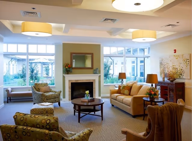 A bright and cozy living room with large windows showing an outdoor garden. The room features a beige sofa with cushions, two patterned armchairs, a round wooden coffee table with a small plant and books, a fireplace with a mirror above it, two table lamps on side tables, and a wooden cabinet with decorative items. The ceiling has recessed lighting and two large circular light fixtures.