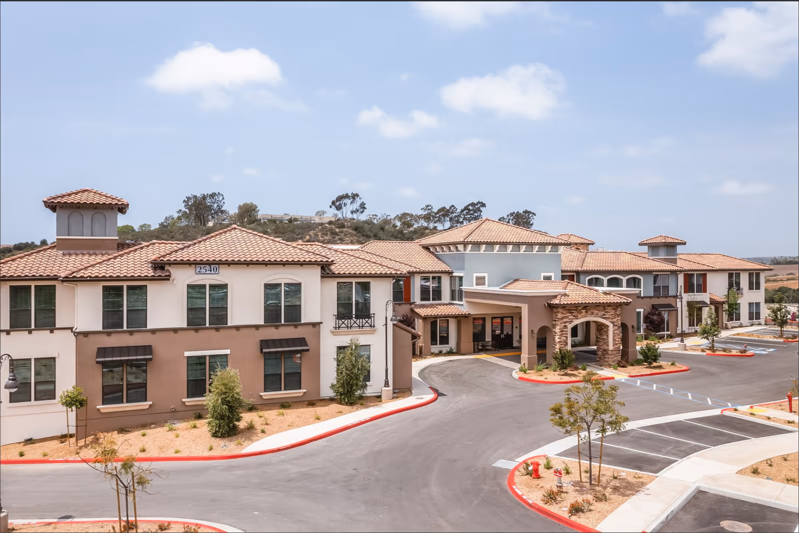 Exterior view of a large, two-story senior living facility with beige and light brown walls, red-tiled roofs, multiple windows, and a covered entrance driveway. The surrounding area includes a paved parking lot, small landscaped areas with young trees and shrubs, and a clear blue sky with some clouds.