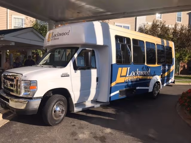 A white and blue shuttle bus parked outside a residential building. The bus has the logo and name 'Lockwood of Clinton' on its side and front. The building in the background has beige siding and multiple windows.