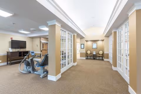 Interior view of a senior living facility hallway with beige walls and carpet. On the left side, there is exercise equipment including a recumbent bike and elliptical machine in front of a wall-mounted TV. The hallway has white-framed glass doors on both sides and two chairs with a small table at the far end.