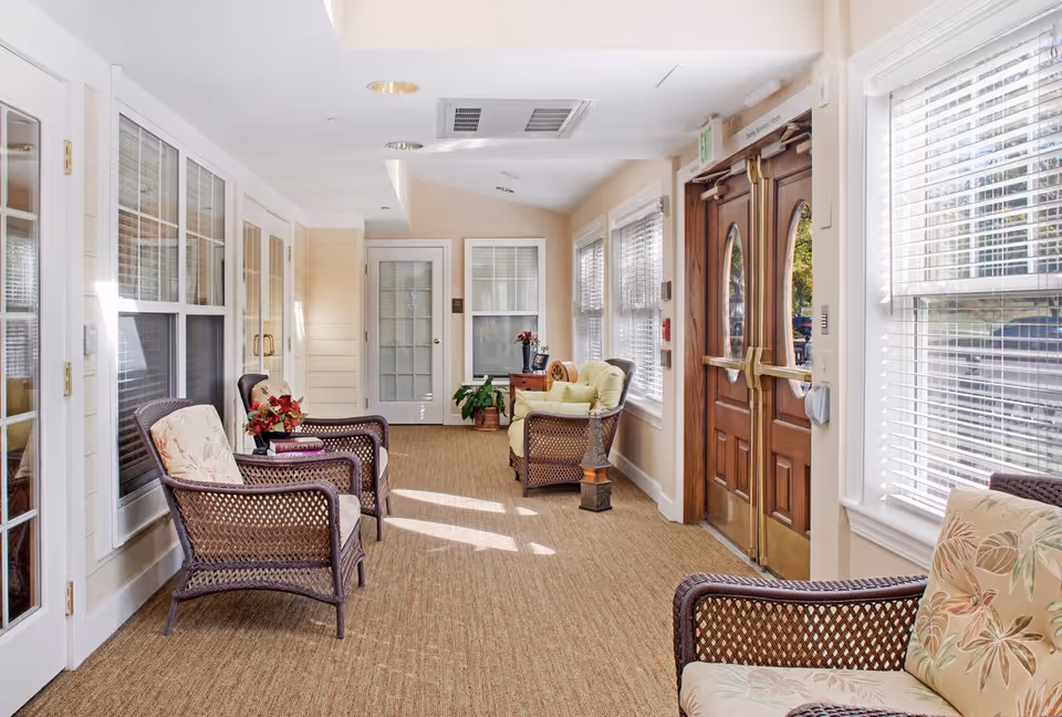 A bright and cozy indoor sitting area with wicker chairs featuring floral cushions, a small wooden table with a plant and flowers, large windows with white blinds letting in natural light, and double wooden doors with glass panels.