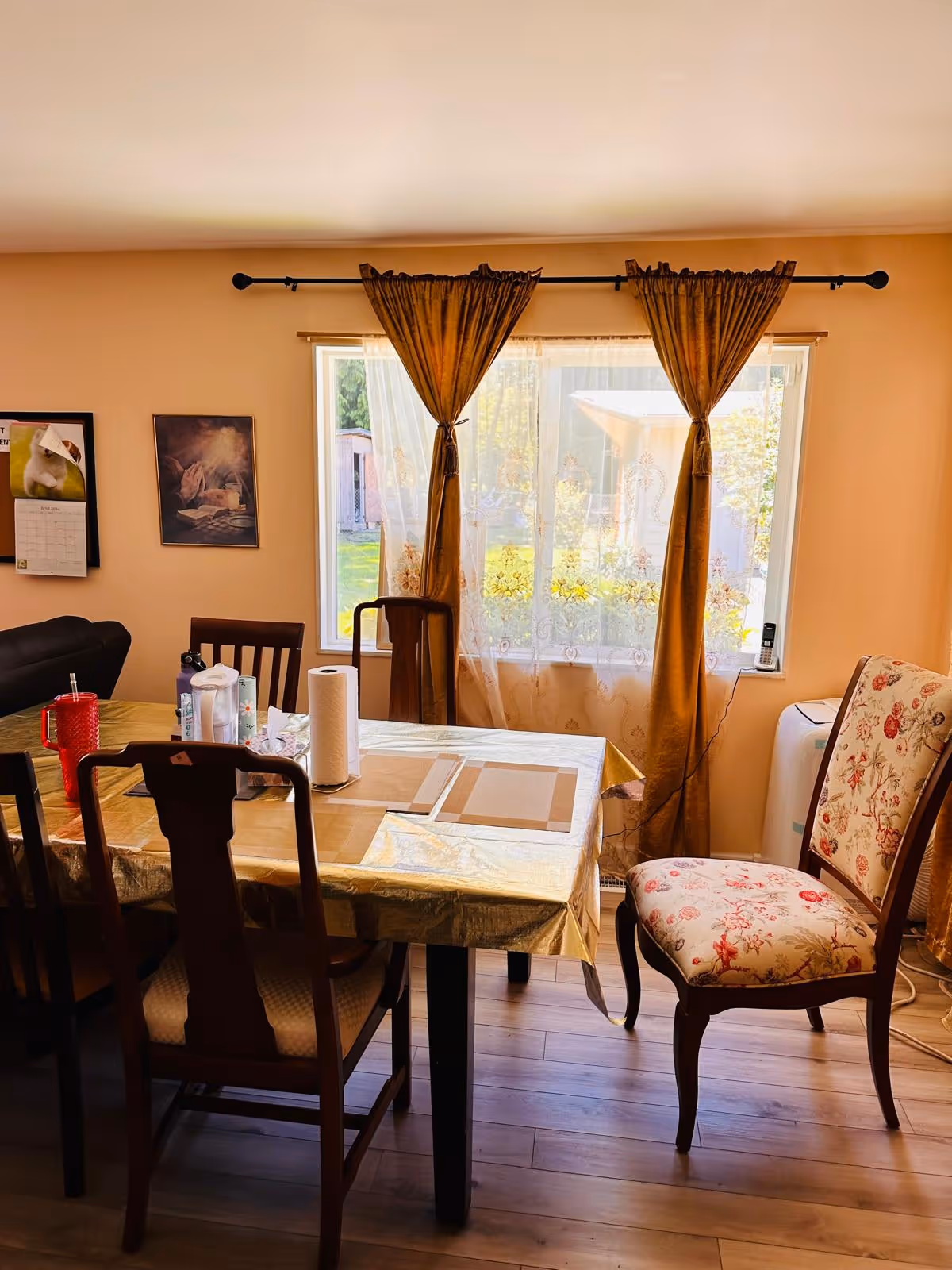 A dining room with a rectangular table covered with a gold tablecloth and placemats. There are four wooden chairs around the table, one of which has floral upholstery. The room has a large window with sheer white curtains and gold drapes tied back, letting in natural light. On the wall, there are two framed pictures and a calendar. The floor is wooden.