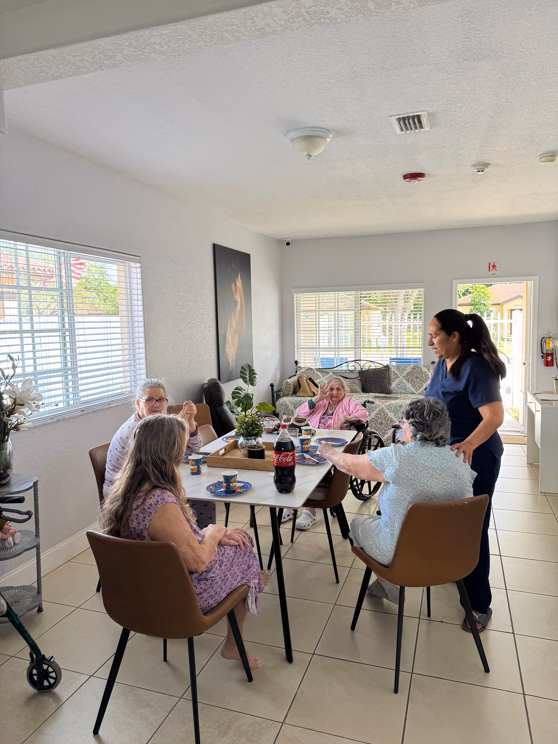 Several elderly residents sit around a dining table while a caregiver stands beside them in a bright common room.
