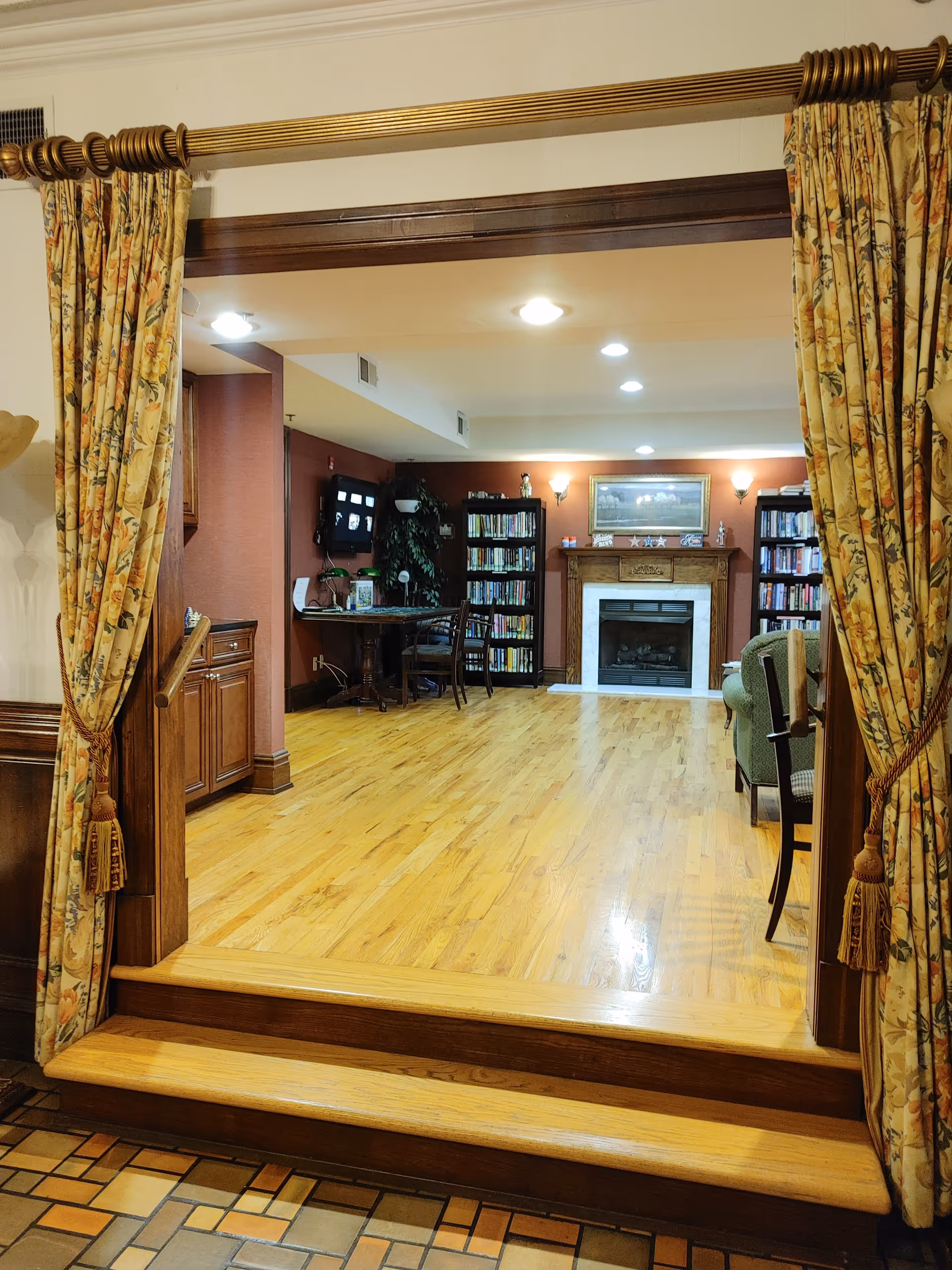 View through an open doorway with floral curtains tied back, leading into a cozy room with wooden floors, bookshelves, a fireplace with a framed painting above it, a table with chairs, and a green armchair.
